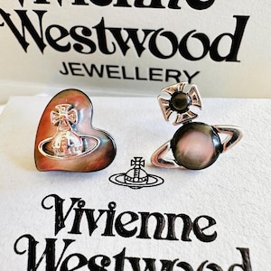 Può includere: Due orecchini Vivienne Westwood con una finitura in madreperla scura e iridescente. Un orecchino è a forma di cuore con un emblema orb argentato, l'altro è un design orb circolare con una croce. Entrambi mostrano il logo del marchio.