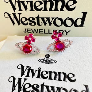Peut inclure: Une paire de boucles d'oreilles à clous argentées avec une pierre ronde fuchsia centrale entourée de cristaux clairs, surmontée d'un motif de fleur fuchsia à quatre pétales. Les boucles sont présentées sur une boîte blanche marquée Vivienne Westwood.