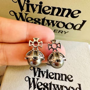 May include: A pair of silver-toned orb earrings featuring a metallic cross emblem atop a spherical grey pearl-like bead. The earrings are displayed against a beige box with the black text Vivienne Westwood printed on the front and top surfaces.