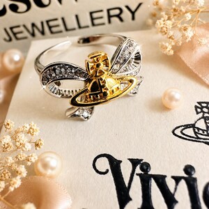 Peut inclure: Une bague argentée ornée d'un emblème central en forme d'orbe doré, entouré d'un nœud argenté incrusté de cristaux transparents. La bague est présentée sur une carte crème portant le texte noir Westwood Jewellery et Vivienne Westwood.