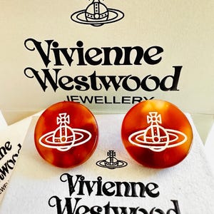 Pode incluir: Um par de brincos redondos com padrão de tartaruga apresentando o logótipo do orbe branco da Vivienne Westwood. Os brincos são exibidos num cartão branco da marca com o logótipo e o texto Vivienne Westwood Jewellery impressos em preto.