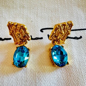 Peut inclure: Une paire de boucles d'oreilles dorées avec une partie supérieure texturée en forme d'éventail et un cristal bleu vif de forme ovale suspendu. Le métal présente une finition organique rugueuse et les pierres bleues sont fixées par des griffes.