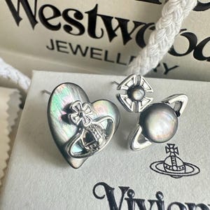 Könnte beinhalten: Zwei silberfarbene Ohrstecker mit dem Vivienne Westwood Orb-Design. Ein Ohrring ist herzförmig mit einer irisierenden dunkelgrauen Einlage, der andere ist eine kreisförmige Kugel mit einem dunkelgrauen sphärischen Zentrum und silbernen Akzenten.