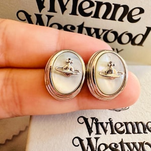 Può includere: Un paio di orecchini ovali color argento con un inserto centrale in madreperla bianca iridescente. Ogni orecchino è decorato con un emblema metallico a forma di globo sormontato da una croce. Il nome Vivienne Westwood è stampato sulla confezione.