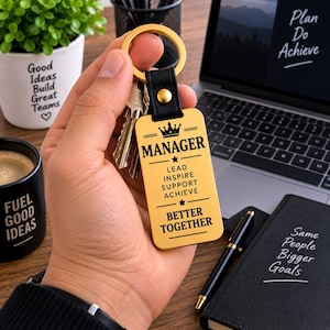 May include: A gold-toned rectangular metal keyring with a black leather strap and a gold rivet. The keyring is engraved with a crown icon, the word MANAGER, and the text LEAD, INSPIRE, SUPPORT, ACHIEVE, and BETTER TOGETHER in black lettering.