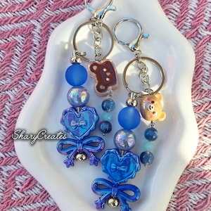 May include: Two keychains featuring blue heart-shaped charms, blue bows, and star accents. Each keychain includes a metal ring, a lobster clasp, and a mix of matte blue, iridescent, and translucent beads. One keychain has a brown bear charm.