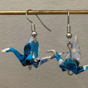 May include: A pair of dangle earrings featuring folded paper crane charms in shades of blue, white, and black with gold-toned accents. The cranes are suspended from silver-toned metal fishhook ear wires, showcasing intricate origami paper patterns.