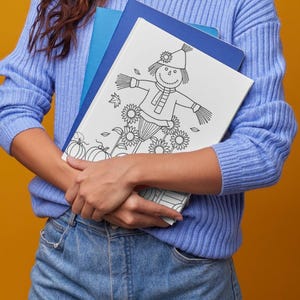 Könnte beinhalten: Ein weißes Malbuch mit einer schwarz-weißen Strichzeichnung einer Vogelscheuche mit Hut und Schal, umgeben von Sonnenblumen und Kürbissen. Das Buch wird vor einem hellblauen Rippenstrickpullover und einer blauen Jeans gehalten.
