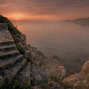May include: A wooden bench with black metal legs sits on a stone patio overlooking a calm sea at sunset. The scene features a stone staircase leading down to the water, surrounded by rocky cliffs under a soft orange and pink sky at dusk.