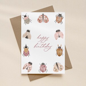 Könnte beinhalten: Eine weiße rechteckige Grußkarte mit dem Text happy birthday in roter Schreibschrift. Die Karte zeigt einen Rand aus acht Aquarellillustrationen verschiedener Insekten und Motten in Rosa-, Braun- und Beigetönen mit floralen Details.