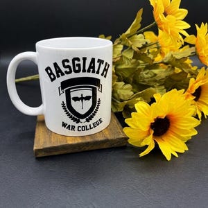May include: White ceramic mug with the text "BASGIATH WAR COLLEGE" in black. The mug features a shield design with a bat and laurel wreath. The mug is on a wooden coaster, with yellow sunflowers in the background.