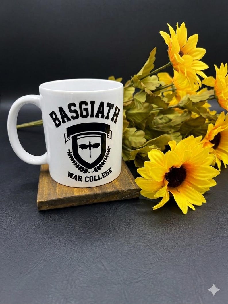May include: White ceramic mug with the text "BASGIATH WAR COLLEGE" in black. The mug features a shield design with a bat and laurel wreath. The mug is on a wooden coaster, with yellow sunflowers in the background.