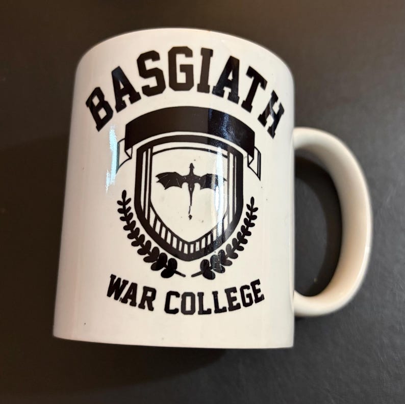 May include: White ceramic mug with the text "BASGIATH WAR COLLEGE" in black. The mug features a crest with a dragon silhouette and laurel leaves. The handle is white.