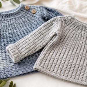 May include: Two long-sleeved sweaters made of crocheted yarn. One sweater features a blue ombre gradient pattern with two small wooden buttons at the neckline. The second sweater is a solid light gray color with a vertical ribbed texture throughout.