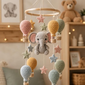 Könnte beinhalten: Ein gehäkeltes Babymobile mit einem grauen Elefanten in der Mitte, umgeben von verschiedenen Heißluftballons und Sternen in Pastelltönen wie Rosa, Gelb, Blau und Grün, die an einem runden Holzrahmen mit weißen Schnüren hängen.