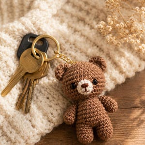 May include: A brown crocheted bear keychain attached to a gold-toned metal ring holding three brass-colored keys. The bear features a small cream-colored muzzle, a dark brown embroidered nose and mouth, and two small black bead eyes.
