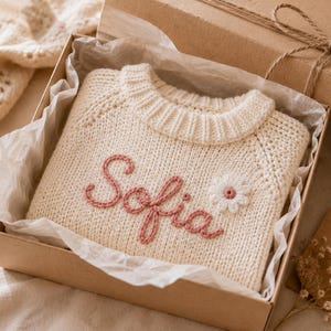 May include: A cream-colored knitted sweater featuring the name Sofia embroidered in pink yarn across the front, accompanied by a small white daisy flower with a pink center, presented inside a cardboard gift box lined with white tissue paper.