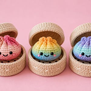 Puede incluir: Tres albóndigas de ganchillo con colores degradados en rosa, amarillo-azul y morado-azul, cada una con ojos de cuentas negras y una sonrisa cosida. Cada albóndiga se asienta dentro de una pequeña cesta de ganchillo beige con tapa.