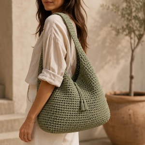 May include: A sage green crochet shoulder bag featuring a textured knit pattern, a wide integrated strap, and a drawstring closure at the top finished with two small matching tassels hanging from the centre of the bag's front panel.