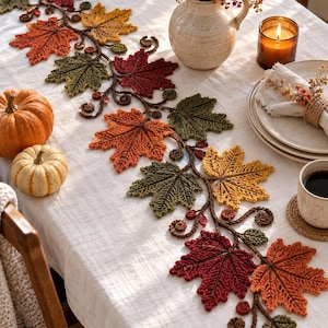 May include: A crochet table runner featuring a vine design with multi-colored autumn leaves in shades of red, orange, yellow, and green. The runner is decorated with small crocheted berries and curled tendrils, laid out on a white tablecloth.