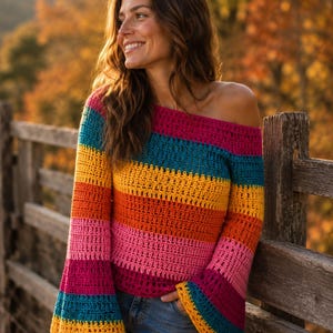 Op de afbeelding: Een gehaakte trui met lange mouwen en horizontale strepen in levendige tinten magenta, talingblauw, geel en oranje. Het kledingstuk heeft een brede, off-shoulder halslijn en uitlopende mouwen, gecombineerd met een klassieke blauwe spijkerbroek.