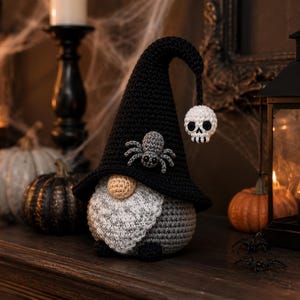 May include: A crocheted gnome figurine featuring a tall black pointed hat adorned with a grey spider and a dangling white skull charm. The gnome has a round beige nose, a textured grey beard, and a rounded grey body with small black feet.