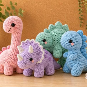 May include: A collection of four crocheted dinosaur toys featuring a pink long-necked dinosaur, a purple triceratops with white horns, a light green stegosaurus, and a blue dinosaur, all with black safety eyes and small pink stitched cheeks.