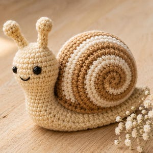 May include: A crocheted snail toy featuring a cream-colored body with two antennae and black plastic eyes. The shell is crafted in a spiral pattern with alternating bands of light brown and cream yarn, creating a textured, rounded appearance.
