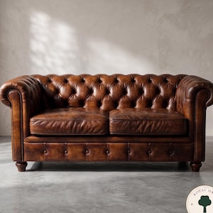 Op de afbeelding: Een bruine leren Chesterfield bank met diepe knoopstoffering op de rugleuning en armleuningen, opgerolde armleuningen, twee zitkussens en een met knopen beklede voorzijde. De bank staat op korte, donkere houten poten en heeft siernagels.