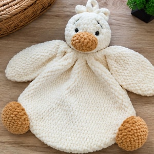 May include: A plush security blanket featuring a crocheted duck head with a white body and head, a tan beak, and two tan feet. The texture is soft and chunky, with black embroidered eyes and a small loop at the top of the head for hanging.