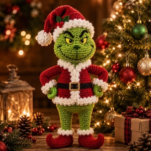Op de afbeelding: Een gehaakte groene wezenpop in een rood-wit kerstmanpak met een zwarte riem en gouden gesp. De pop heeft een puntige rode muts met een witte pompon en witte rand, en draagt rode schoenen met omhooggekrulde punten en een gestikt gezicht.