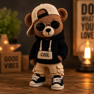 Op de afbeelding: Een bruine gehaakte teddybeer met een beige honkbalpet, ronde zwarte zonnebril, zwarte hoodie met het woord COOL op een rechthoekige patch, beige cargobroek en zwart-witte sneakers met witte veters.