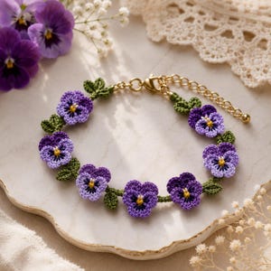 Può includere: Un bracciale caratterizzato da una serie di fiori di viola all'uncinetto nei toni del viola e del violetto chiaro con centri gialli e piccole foglie verdi, collegati da una catena in metallo dorato con chiusura a moschettone e prolunga.