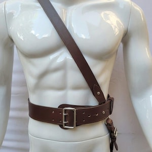 May include: A dark brown leather belt with a silver-toned metal buckle and a matching diagonal shoulder strap. The belt features multiple adjustment holes and is worn across the torso, connecting to the shoulder strap with a metal loop and rivets.