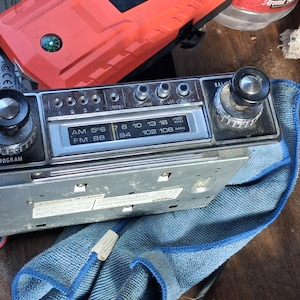 May include: A vintage automotive AM/FM radio receiver featuring a silver faceplate with black knobs, push buttons, and a frequency display showing AM 556 to 1600 kHz and FM 88 to 108 MHz. The unit has a metallic chassis and is resting on a blue cloth.