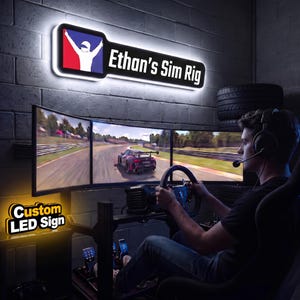 May include: A rectangular wall-mounted LED sign featuring a black frame with white backlighting. The sign displays a graphic of a white racing driver silhouette on a blue and red background, followed by the white text "Ethan's Sim Rig".