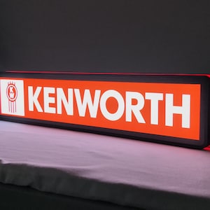 Op de afbeelding: Een rechthoekig LED-bord met het Kenworth-logo en de merknaam in witte letters op een feloranje achtergrond. Het bord heeft een zwarte omlijsting met een gloeiende rode lichtstrip aan de zijkant voor een heldere verlichte weergave.