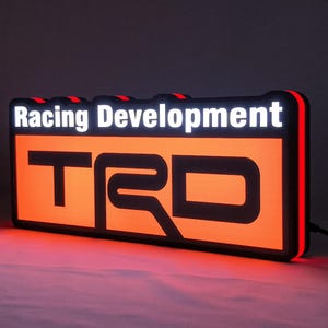 Op de afbeelding: Een rechthoekig LED-bord met de tekst Racing Development in witte letters boven het zwarte TRD-logo. Het bord heeft een fel verlichte oranje achtergrond met een zwarte rand en een rode lichtstrip langs de zijkant.