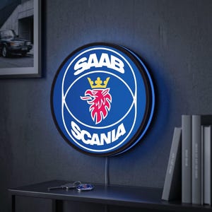 May include: A circular wall-mounted LED sign featuring the blue, white, and red Saab Scania logo with a central griffin emblem wearing a yellow crown. The sign emits a soft blue glow from behind its circular frame, displaying the text SAAB SCANIA.