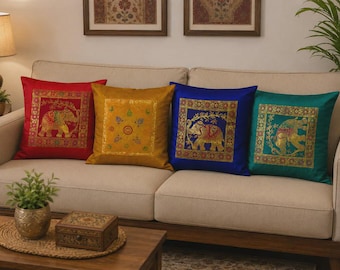 Op de afbeelding: Een set van vier vierkante sierkussens in rood, goud, blauw en groenblauw. Elk kussen heeft een centraal vierkant paneel met gedetailleerd goudborduursel. Twee kussens tonen een olifantenmotief, één een bloemenpatroon en één een mandala-ontwerp.