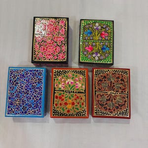 May include: Five rectangular boxes with lids, each featuring distinct hand-painted floral patterns. The designs include pink flowers on black, colorful blossoms on green, blue flowers on blue, and multi-colored floral motifs on red and black backgrounds.