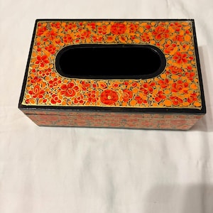 May include: A rectangular tissue box cover featuring a black base with a vibrant hand-painted floral pattern in shades of orange, red, and yellow. The top surface has a central oval opening, and the sides are decorated with a dense, repeating botanical design.