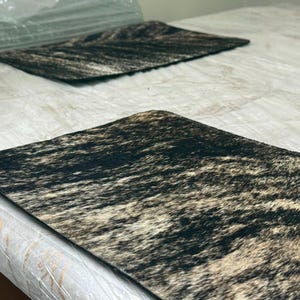 Pode incluir: Dois jogos americanos retangulares com um padrão texturizado inspirado em pele de animal em tons de preto, castanho escuro e creme. Os jogos americanos têm um design plano com cantos arredondados e uma estampa orgânica consistente.