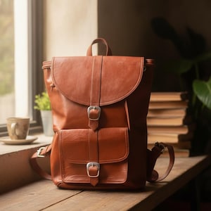 Puede incluir: Una mochila de cuero color coñac que presenta una solapa superior grande con un cierre de hebilla de metal plateado, un bolsillo frontal con una hebilla a juego, un asa superior y correas ajustables con herrajes de metal plateado.