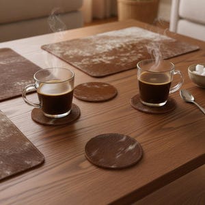 May include: Two clear glass mugs filled with steaming dark coffee, each resting on a round brown textured coaster. Three additional round brown coasters are arranged on the wooden surface, alongside two rectangular brown placemats with white mottled patterns.