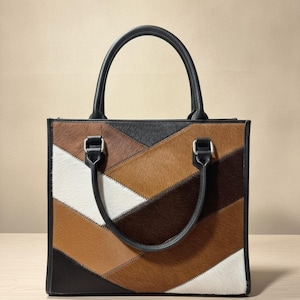 May include: A square-shaped handbag featuring a patchwork design of cowhide panels in shades of white, tan, brown, and black. The bag includes two black leather top handles attached with silver-toned metal hardware and black leather trim along the edges.