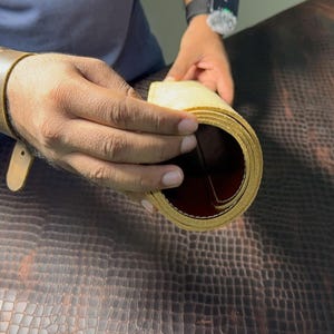 Op de afbeelding: Een opgerold stuk lichtbruin leer met een gladde, donkerroodbruine binnenzijde. Het leer wordt in een cilindrische vorm gehouden, wat het contrast laat zien tussen de lichte buitenkant en de donkere, afgewerkte binnenkant van het materiaal.