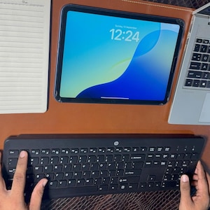 Könnte beinhalten: Eine schwarze kabellose Tastatur mit HP-Logo, ein Tablet mit der Anzeige 12:24, ein silberner Laptop mit sichtbarer Tastatur und Touchpad sowie ein offenes Notizbuch mit linierten Seiten, angeordnet auf einer glatten braunen Lederschreibtischunterlage.