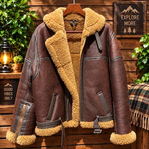 May include: A dark brown leather jacket featuring a thick tan shearling lining and collar. The jacket includes a front zipper closure, two side pockets, adjustable buckled straps at the hem, and zippered cuffs with tan shearling trim at the sleeves.