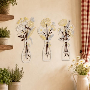Op de afbeelding: Drie wanddecoraties met gestileerde bloemstukken in glazen flessen. Elk stuk toont witte, gele en bruine bloemen met stelen. De middelste en rechter vaas zijn voorzien van de teksten "LOVE" en "YOU" op de hals van de flessen.